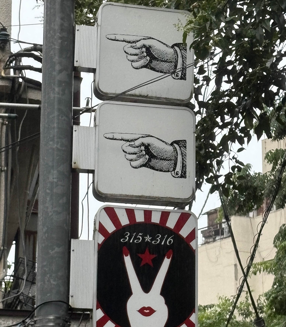 three signs, top 2 are a hand pointing left, third is a peace sign with a mouth on the hand